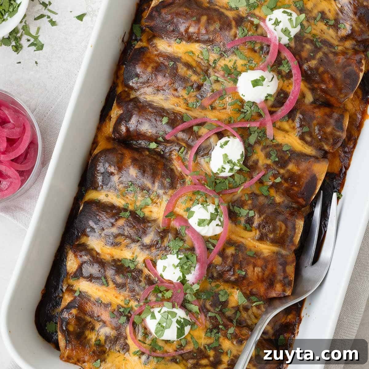 A baking dish full of shredded beef enchiladas with a deep red enchilada sauce cilantro, and pickled onions.