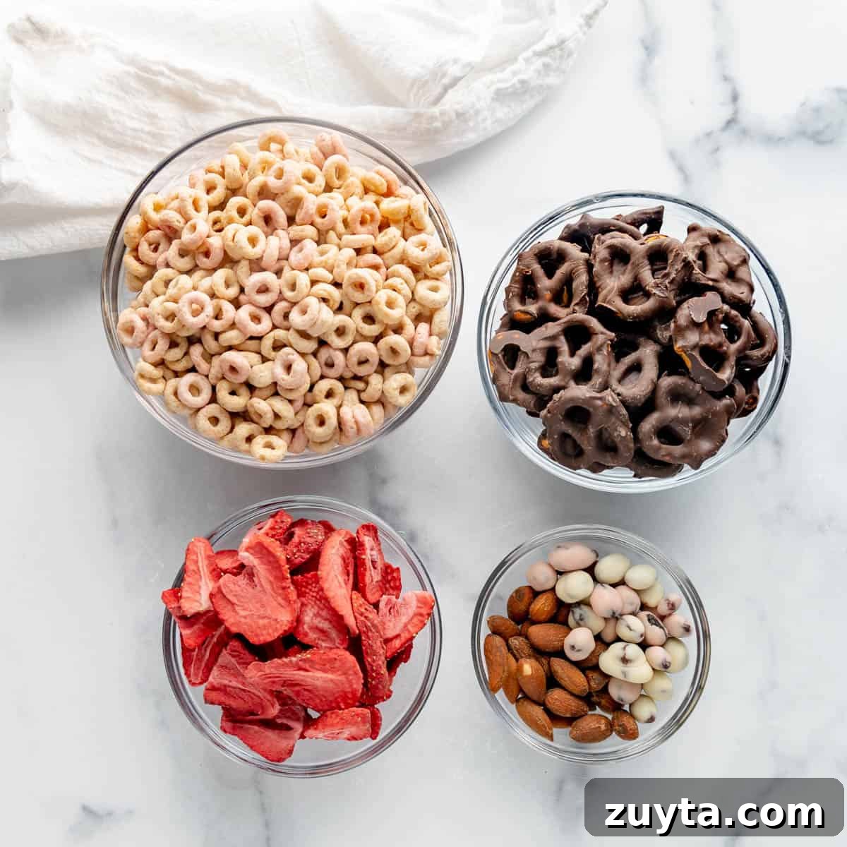 Valentine's Snack Mix Recipe with Chocolate and Pretzels 2 Four glass bowls filled with cereal, chocolate-covered pretzels, freeze-dried strawberries, and mixed nuts on a marble surface. A white cloth is partially visible.