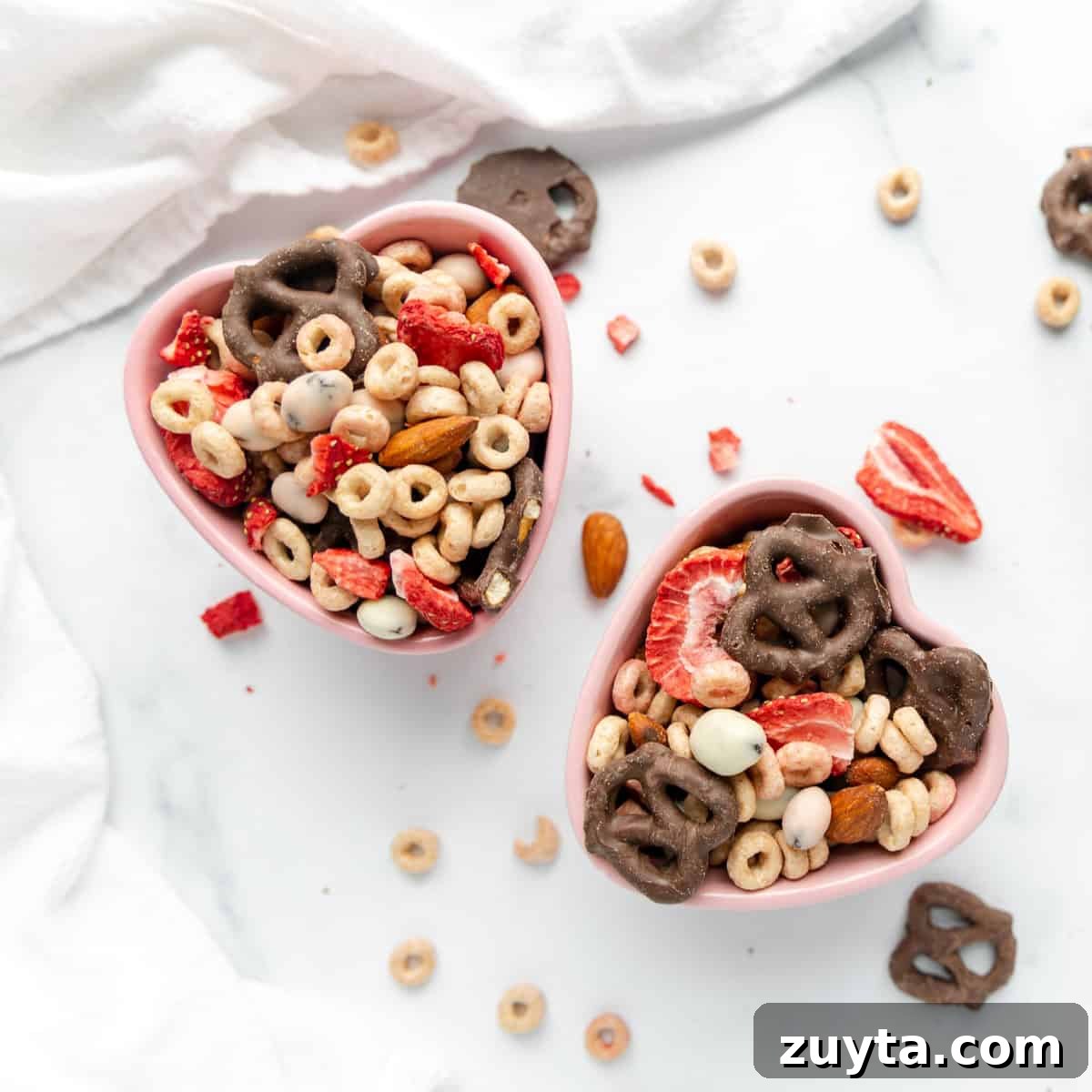 Valentine's Snack Mix Recipe with Chocolate and Pretzels 3 Two heart-shaped bowls filled with cereal, pretzels, almonds, and dried strawberries on a white surface with scattered ingredients.