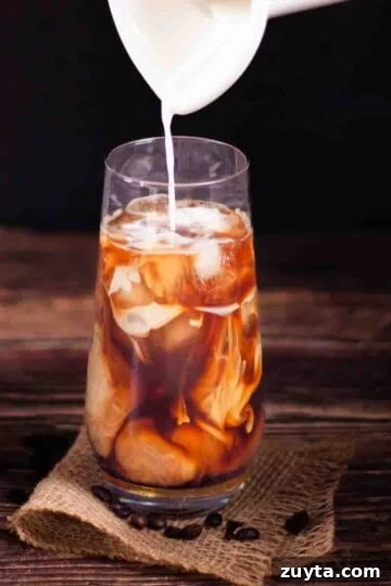 Cream being poured over coffee and ice cubes in a tall glass to make the perfect iced coffee at home.