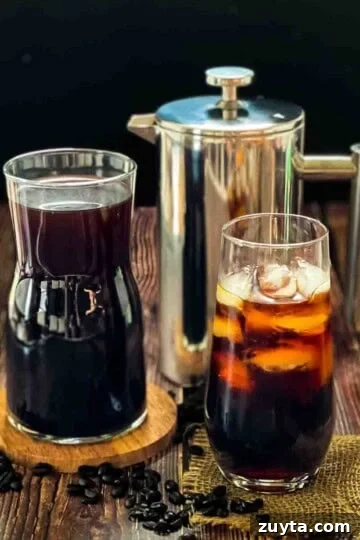 Cold brew coffee in a tall glass with ice, with a pitcher filled with cold brew and a French press coffee maker behind it.