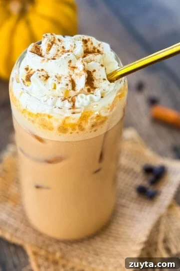 Glass of homemade iced pumpkin spice latte with ice cubes and whipped cream, surrounded by coffee beans and cinnamon sticks.