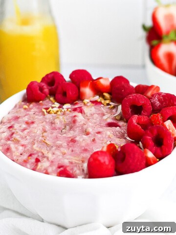 Pink raspberry oatmeal in a white bowl, garnished with fresh raspberries and strawberries.