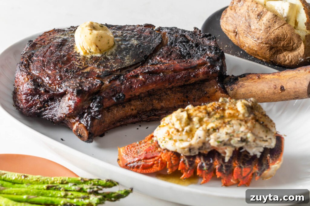 Tomahawk Steak Recipe: Grill and Reverse-Sear 3 cooked tomahawk steak topped with a scoop of compound butter on a plate with a cooked lobster tail. asparagus spears and a baked potato with butter in the background