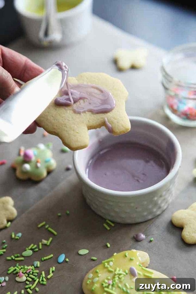 Lemon Tea Cakes Recipe with Zesty Glaze 2 a knife drizzles purple frosting onto a sheep-shaped cookie held in hand above a countertop lined with parchment paper