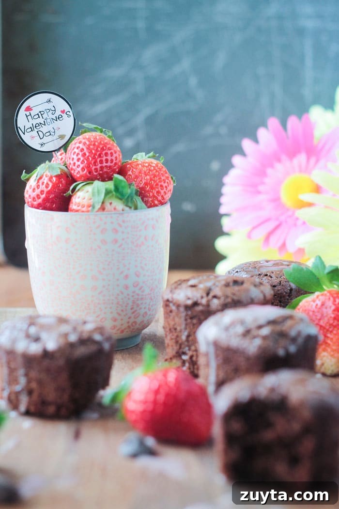 Tahini Chocolate Chip Cupcakes - These adorable little mini cupcakes are perfect for Valentine's Day. Treat your sweetie...or your kids...or yourself...to some chocolate indulgence made healthier with no dairy, no eggs, no oil, and no refined sugar. A small pink cup of fresh strawberries, with a Happy Valentine's Day cupcake topper sticking out of the top, surrounded by tahini chocolate chip cupcakes. Pink flowers in the background.