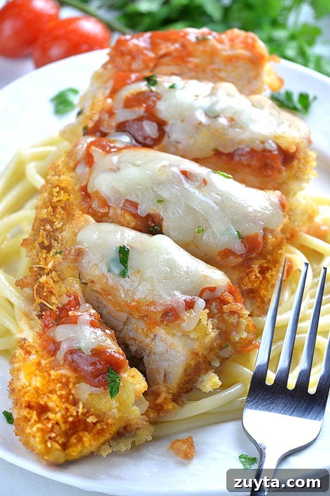 Crispy Baked Chicken Parmesan Recipe with Marinara 2 Whole piece of Baked Chicken Parmesan on a white plate laying on spaghetti with fork.