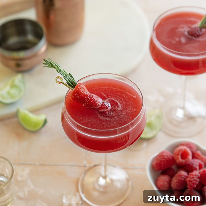 two glasses of a raspberry cosmopolitan next to a bowl of raspberries and slices of lime